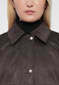 Close-up van een persoon in een donkerbruine suède jas met een brede kraag, zilveren drukknoopjes en een zwart coltrui eronder.