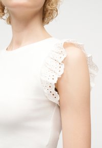 Femme portant un haut blanc sans manches avec des détails en dentelle ajourée à volants sur l'épaule, sur un fond uni clair.