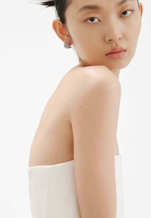 Modelo que lleva una blusa blanca sin mangas con una textura suave, mostrando un pendiente plateado tejido de forma circular y con patrones sutiles.