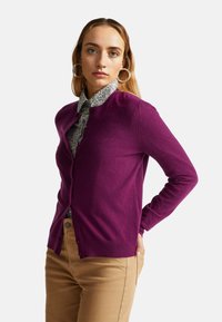 Fuchsiový kardigan vyrobený z měkké látky, s kulatým výstřihem a zapínáním na knoflíky, vrstvený přes vzorovanou košili, doplněný béžovými kalhotami.