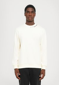 Man with short hair wearing a white sweatshirt with "EA7 Emporio Armani" text near the collar and black pants, standing against a plain background.