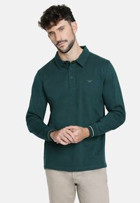 Polo de manga larga verde oscuro hecho de una tela suave. Presenta un cuello con solapa, una tapeta de tres botones y detalles en contraste en los puños.