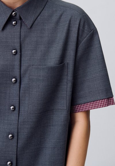 Camicia a bottoni in tessuto grigio scuro con taschino sul petto, bottoni argentati lucidi e motivo a quadri rossi sui polsini. Tessuto liscio.
