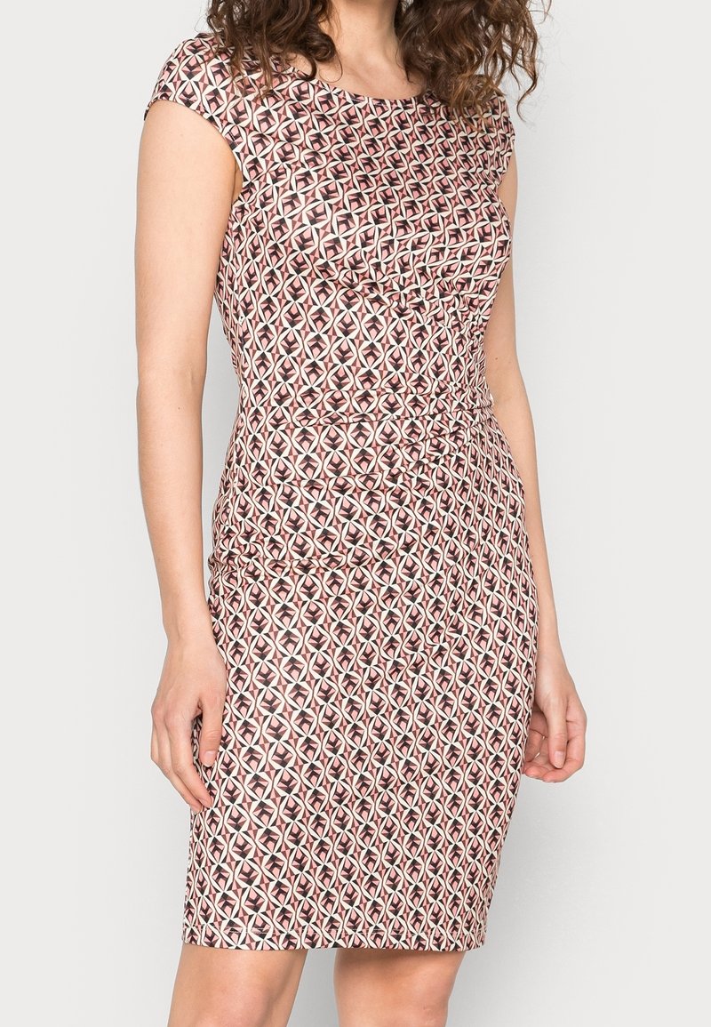 Femme portant une robe ajustée longueur genoux avec des manches courtes et un motif géométrique répétitif en rose, noir et blanc.
