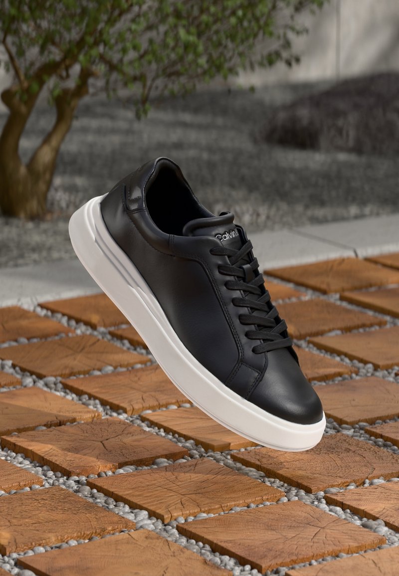 Baskets en cuir noir à semelle blanche et lacets noirs flottant au-dessus d'un sol extérieur en dalles de bois et galets, avec un arbre en arrière-plan.