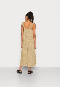 Robe longue en tissu jaune ornée d'un motif floral. Dotée de bretelles à volants et d'une coupe ample et fluide. Portée avec des sandales noires.