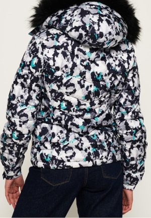 Osoba nosząca białą, czarną i zielononiebieską pikowaną kurtkę z kapturem wykończonym czarnym futrem, w połączeniu z ciemnoniebieskimi jeansami.