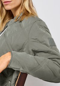 Světle šedá prošívaná bomber bunda s texturovaným švem, žebrovanými manžetami a logem na rukávu. Hladká tkanina s matným povrchem.