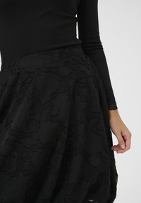 Ženska v črni bluzi z dolgimi rokavi in črni krilu visoko v pasu s teksturiranim cvetličnim vzorcem, roka počiva ob robu krila.