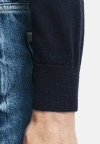Marineblauer Strickpullover mit Rippbund an den Ärmeln. Verfügt über dezentes Branding am Rand, kombiniert mit hellblauen, stark verwaschenen Jeans.