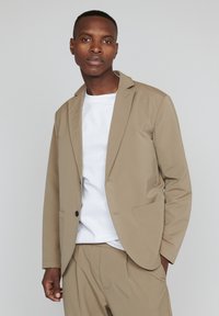 Šviesiai beige spalvos švarkas su vienu juodu mygtuku ir dviem priekinėmis kišenėmis, dėvimas virš balto marškinio, pasižymintis moderniu, pritaikytu siluetu.