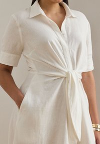 Balta lina kleita ar īsām piedurknēm, apkakli un sasienamu jostas detaļu. Mīksta tekstūra, kabatas un brīvs piegriezums.