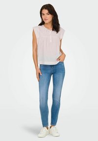 Jasnoróżowa bezrękawnikowa bluzka z zapięciem na guziki i marszczonymi detalami, zestawiona z niebieskimi wąskimi dżinsami i białymi trampkami. Gładki materiał.