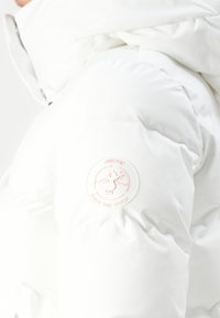 Witte pufferjack met een gladde, gewatteerde stof en een rond wit en perzikkleurig logo op de mouw. Het design is minimalistisch en functioneel.