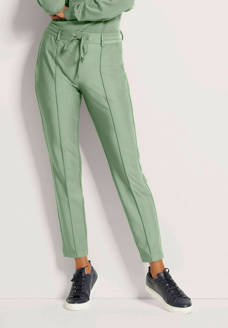 Lichtgroene tapered broek met een trekkoord in de taille, met contrasterende zijnaad en gecombineerd met donkerblauwe sneakers. Het stof heeft een gladde textuur.