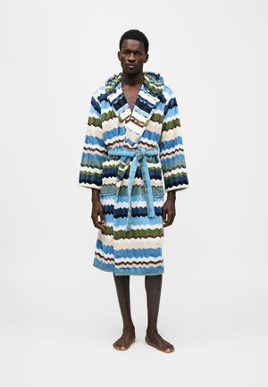 Un homme se tient pieds nus devant un fond uni et présente une robe de chambre à motifs en zigzag bleu, vert et blanc, nouée avec une ceinture.