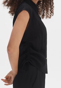 Černá halenka bez rukávů s texturovaným povrchem, s límečkem, bočními řasenými detaily a zlatými knoflíky. Zobrazeno z bočního úhlu.