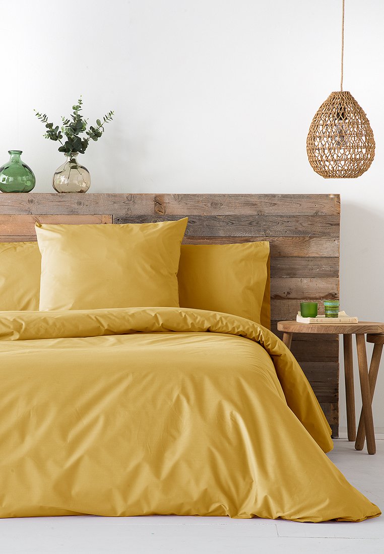 Gelbe Bettwäsche mit glatter Textur, kombiniert mit zwei passenden Kissen auf einem Holzbetthead. Einfache Rattan-Pendelleuchte und dekorative Vasen in der Nähe.