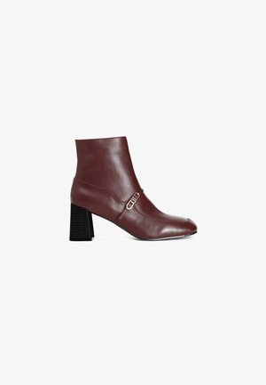Burgundy læder ankelstøvle med en kraftig sort hæl og en dekorativ spænde detalje. Har en spids tå og glat tekstur.