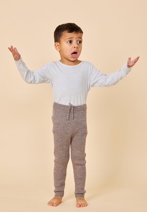 Jeune garçon debout pieds nus, bras levés, bouche ouverte, portant un t-shirt rayé à manches longues et un pantalon en maille gris sur un fond beige.