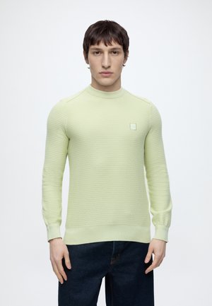 Mladý muž s krátkymi hnedými vlasmi, oblečený v svetlozelenom textúrovanom svetri s dlhým rukávom a tmavomodrých džínsoch, stojaci pred jednoduchým bielym pozadím.