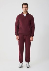 Sweat-shirt bordeaux zippé avec une poche frontale, assorti à un pantalon de jogging. Présente des poignets côtelés et une texture lisse. Baskets blanches.