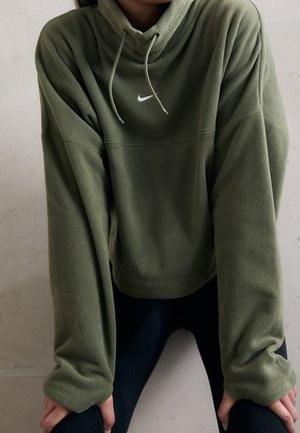 Osoba w za dużej oliwkowozielonej bluzie z kapturem Nike z polaru z troczkami, pochylona do przodu z rękami opartymi na kolanach na jednolitym tle.