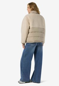Kobieta odwrócona plecami, w beżowej kurtce sherpa i pikowanej, w szerokich niebieskich dżinsach i szarych trampkach na białym tle.