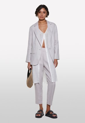 Costume en lin gris clair avec un long blazer et un pantalon fuselé, associé à une chemise blanche à boutons et des sandales noires. Accentué par un sac en osier.