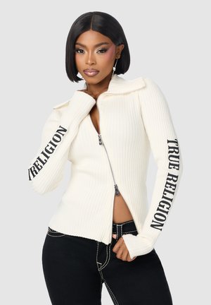 Vrouw met kort zwart haar die een witte zip-up trui draagt met de tekst "TRUE RELIGION" op de mouwen en een zwarte broek met witte stiksels.