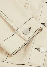 Beige denim-kangasta, jossa on kontrastoiva musta ommel, rullatut hihat ja hopeiset nepparilukot.