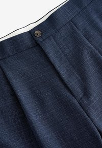 Marineblaues Hose mit strukturiertem Stoff, ausgestattet mit einem Knopfverschluss und einer gefalteten Vorderseite. Weiße Fütterung ist am Bund sichtbar.