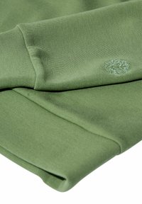 Tejido textil verde con textura suave, que presenta una manga plegada y un sutil logo bordado en la esquina superior.