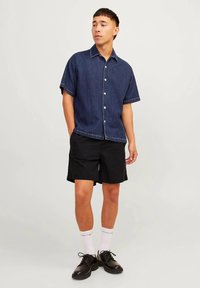 Camisa de mezclilla oscura de manga corta con botones, costuras blancas, combinada con pantalones cortos negros y zapatos negros. Calcetines blancos visibles. Fondo neutro.