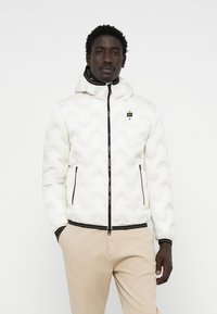 Witte pufferjack met een chevronpatroon, ritssluiting, zwarte afwerking en een capuchon. Heeft zijzakken en een merklogo op de borst.