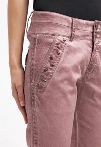 Rosa byxor med en lätt glänsande yta, som har stjärnformade nitar och knappdetaljer på framsidan. Slät textur och åtsittande design.