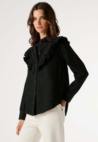 Čierna rifle košeľa s výrazným volánovým golierom, zdobená malými cvokmi, s dlhými rukávmi a zaobleným lemom, v kombinácii s bielymi nohavicami.