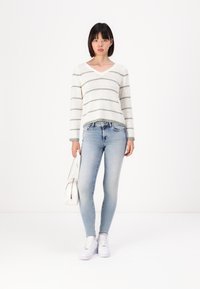 Hvit genser med grønne striper, V-hals og lange ermer, kombinert med lyseblå skinny jeans og hvite joggesko. Modell holder en hvit veske.