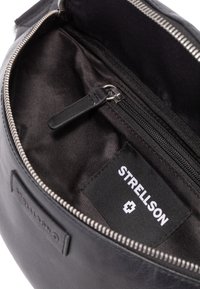 Sort læderpung med en sølvfarvet lynlås. Indersiden har en glat stofforing og en label med teksten "STRELLSON." Indeholder en lynlåslomme.