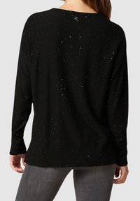 Schwarzer Pullover mit lockerer Passform, der eine strukturierte Oberfläche mit verstreuten Funkelndetails, gerippten Bündchen und einem Knopfdetail am Rücken aufweist.