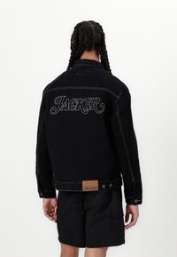 Schwarze Jeansjacke mit weißem Nähen, "JACKER" auf dem Rücken gestickt und einem braunen Lederetikett am Saum. Klassisches Design mit Knopfverschluss.