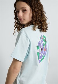 Lyseblå bomulds t-shirt med et farverigt cirkulært grafisk motiv på ryggen, der viser illustrerede dyr og planter. Korte ærmer, afslappet pasform.