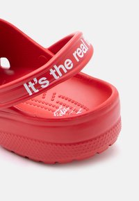 Κοντινή λήψη κόκκινου πλαστικού σανδαλιού με υφασμένο πέλμα και εν μέρει λευκή επιγραφή στο λουρί που λέει «Είναι το πραγματικό t.»