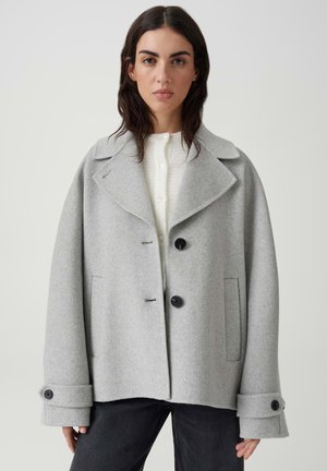 Donna che indossa un cappotto di lana grigio chiaro con grandi rever, bottoni neri, cinturini ai polsini e tasche laterali sopra una maglia bianca e pantaloni neri.