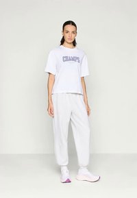 Witte katoenen t-shirt met de tekst "CHAMPS" in paars, gecombineerd met lichtgrijze joggingsbroek en witte sneakers met roze accenten.