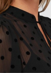 Blouse noire transparente à pois, avec un col haut et des boutons au niveau du cou. Texture douce avec des manches bouffantes.