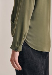 Blusa verde oliva con maniche lunghe, polsini bottonati e una texture liscia, abbinata a pantaloni in denim scuro. Vestibilità rilassata con fessure laterali.