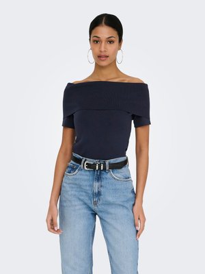 Ženska v temnomornem topu z odkrito ramo, svetlo modrih kavbojkah z visokim pasom, črnem pasu in velikih uhanih v obliki obroča, stoji ob enobarvnem ozadju.