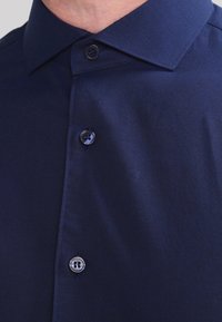 Navyblauwe overhemdd met een puntige kraag en drie zichtbare knopen aan de voorkant op de bovenste torso van een persoon.