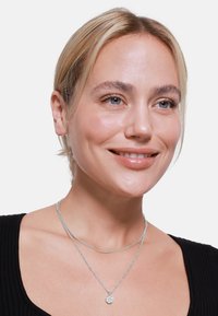 Zilveren gelaagde necklaces met een getextureerde hanger aan de onderste ketting, gedragen over een zwart geribd topje. Glad metalen oppervlak en slank design.